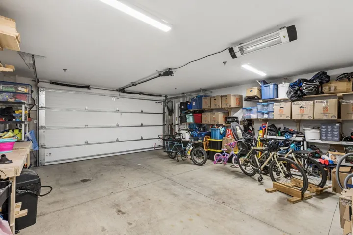 Spacious garage featuring a sectional overhead door, exposed ceiling joists, durable concrete flooring, fluorescent lighting, and extensive wall-mounted shelving