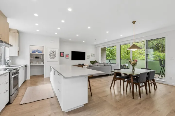 Open-concept living space featuring wood-finish flooring throughout, a modern kitchen island with a light-colored countertop, and a dining area illuminated by a contemporary pendant light