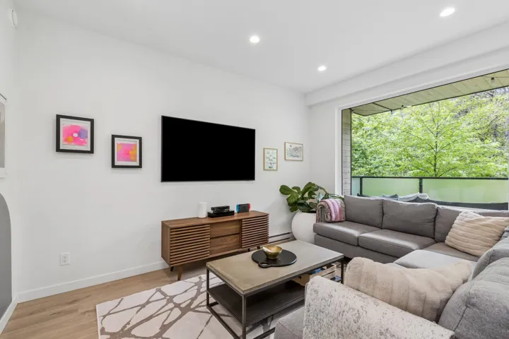 Bright living area featuring wood-finish flooring, a contemporary wood media console, recessed lighting, and a wide sliding glass door opening to a balcony with a glass railing