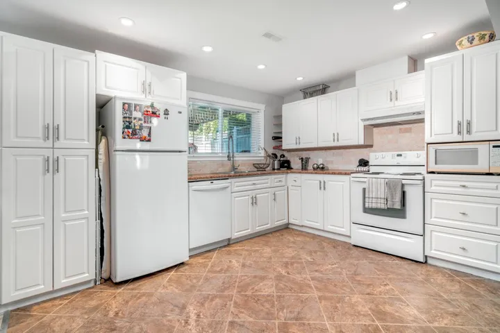 Kitchen featuring white appliances, under cabinet range hood, backsplash, white cabinetry, and recessed lighting