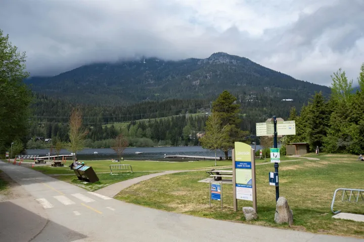 Lush green landscape featuring a paved pathway, mature trees, a distant lake, and a mountainous backdrop