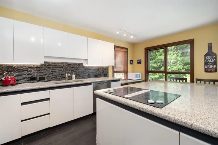 Kitchen featuring white flat-panel cabinetry, a speckled countertop, dark mosaic tile backsplash with under-cabinet lighting, and an integrated cooktop on the island