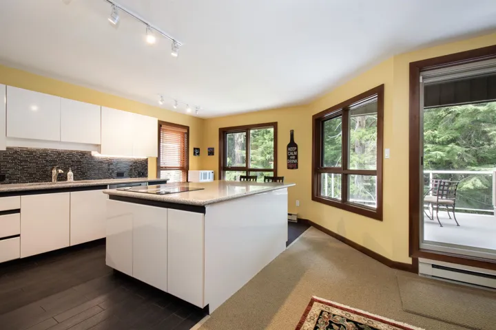 Kitchen featuring white high-gloss cabinetry, dark wood-finish flooring, a central island with an integrated cooktop, a mosaic tile backsplash, and multiple windows with dark wood trim
