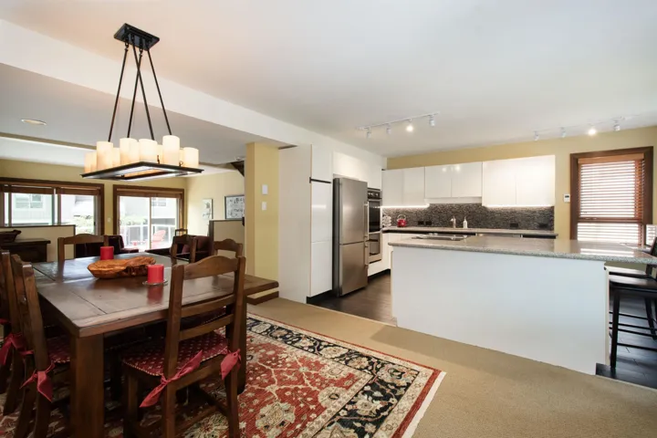 Open-concept dining area featuring a rectangular chandelier, wood-finish flooring, and a kitchen with white cabinetry, stainless steel appliances, and a central island with a stone-finish countertop