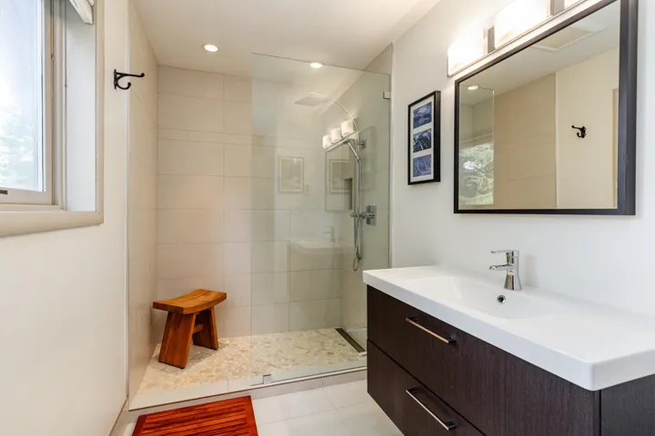 Bathroom featuring a floating dark wood-finish vanity with an integrated white sink, a frameless glass-enclosed shower with pebble-tile flooring, and a wall-mounted mirror with overhead lighting