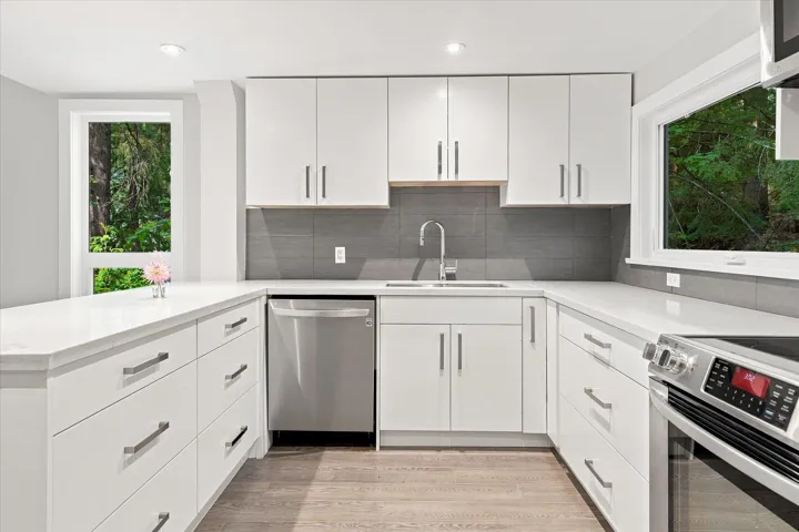 U-shaped kitchen featuring white cabinetry, light-toned countertops, a stainless steel dishwasher, a gray tile backsplash, and light wood-finish flooring