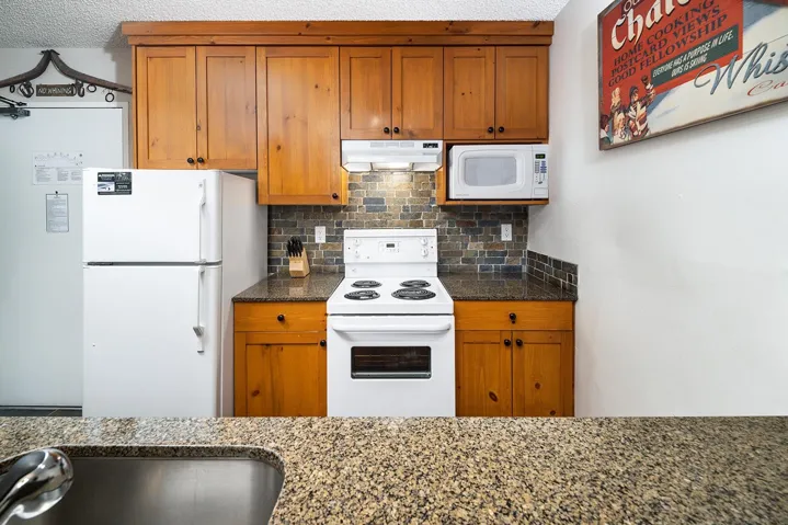 Kitchen featuring white appliances, decorative backsplash, dark stone counters, and wood finish cabinetry