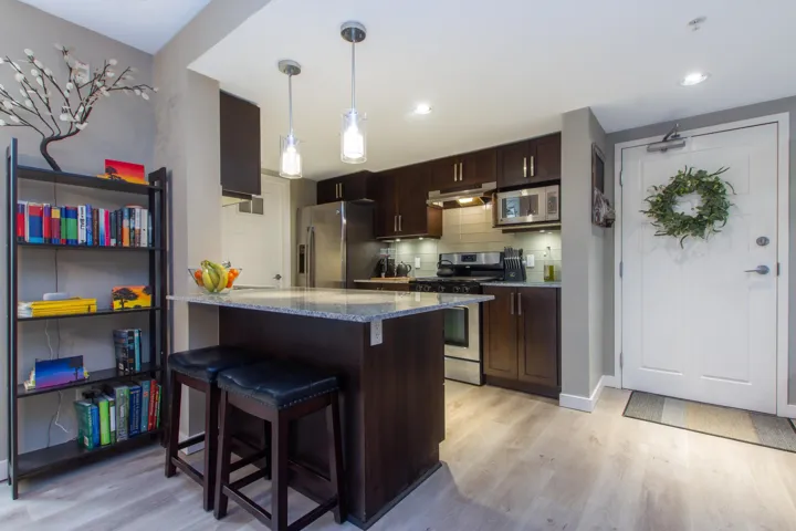Kitchen featuring light wood finished floors, stainless steel appliances, dark brown cabinetry, under cabinet range hood, and tasteful backsplash