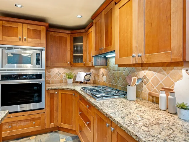 Kitchen with appliances with stainless steel finishes, brown cabinets, tasteful backsplash, light stone countertops, and glass insert cabinets
