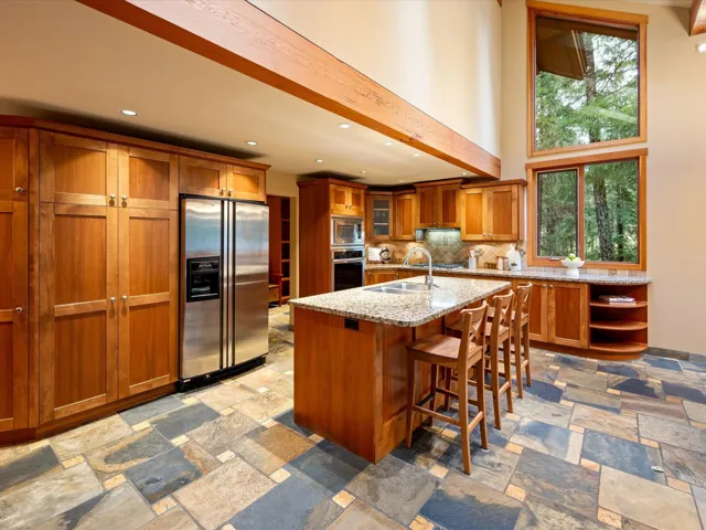 Kitchen with brown cabinetry, appliances with stainless steel finishes, light stone counters, glass insert cabinets, and a kitchen bar
