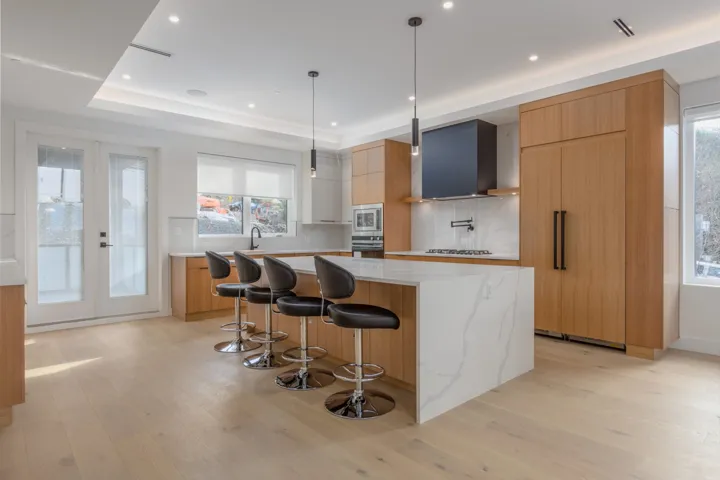 Contemporary kitchen featuring a waterfall island with seating for four, light wood-finish cabinetry, integrated paneled refrigerator, built-in cooktop with a black range hood, and light wood-finish flooring