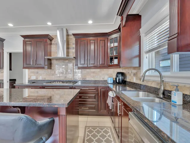 Kitchen featuring dark wood cabinetry, granite countertops, a tiled backsplash, a stainless steel range hood, and recessed lighting