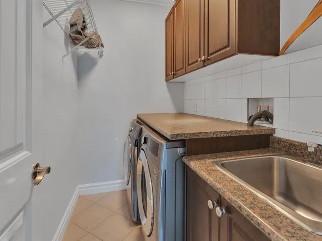 Dedicated laundry space featuring a stainless steel utility sink, granite-style countertops, wood-finish cabinetry, white tile backsplash, and ceramic tile flooring