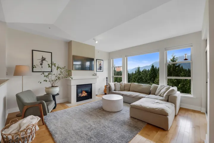 Living room with ample natural light, a gas fireplace with a mantel, light wood-finish flooring, and large windows showcasing distant mountains and evergreen trees