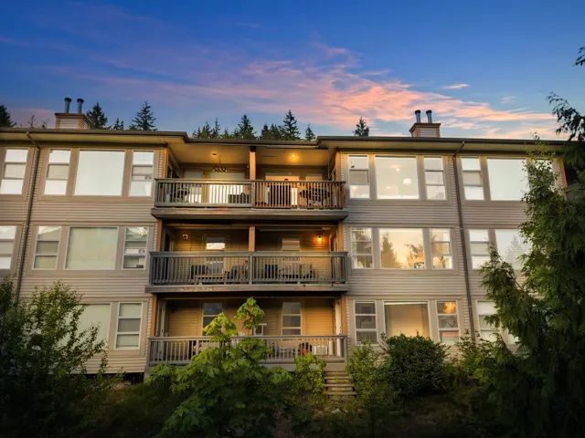 Multi-level residential building featuring multiple private balconies with wood railings, extensive window placements, and a natural tree-lined backdrop