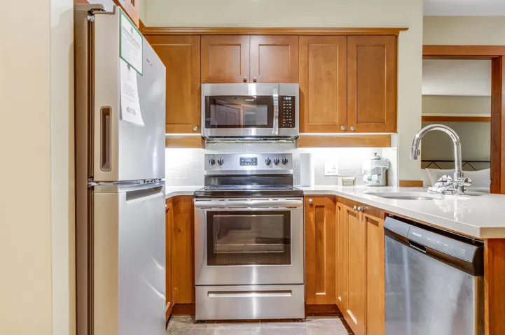 Kitchen with brown cabinetry, stainless steel appliances, decorative backsplash, light stone counters, and a peninsula