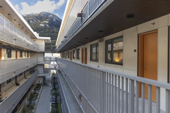 Modern multi-story building featuring exterior corridors with light gray railings, contrasting beige wall panels, and warm-toned entry doors
