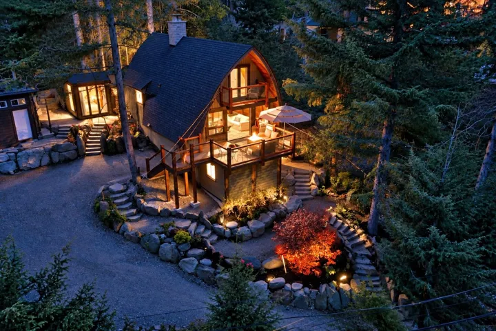 Gable roofline with stone chimney, multiple outdoor decks, terraced landscaping with stone retaining walls, gravel driveway, and a detached structure with large windows