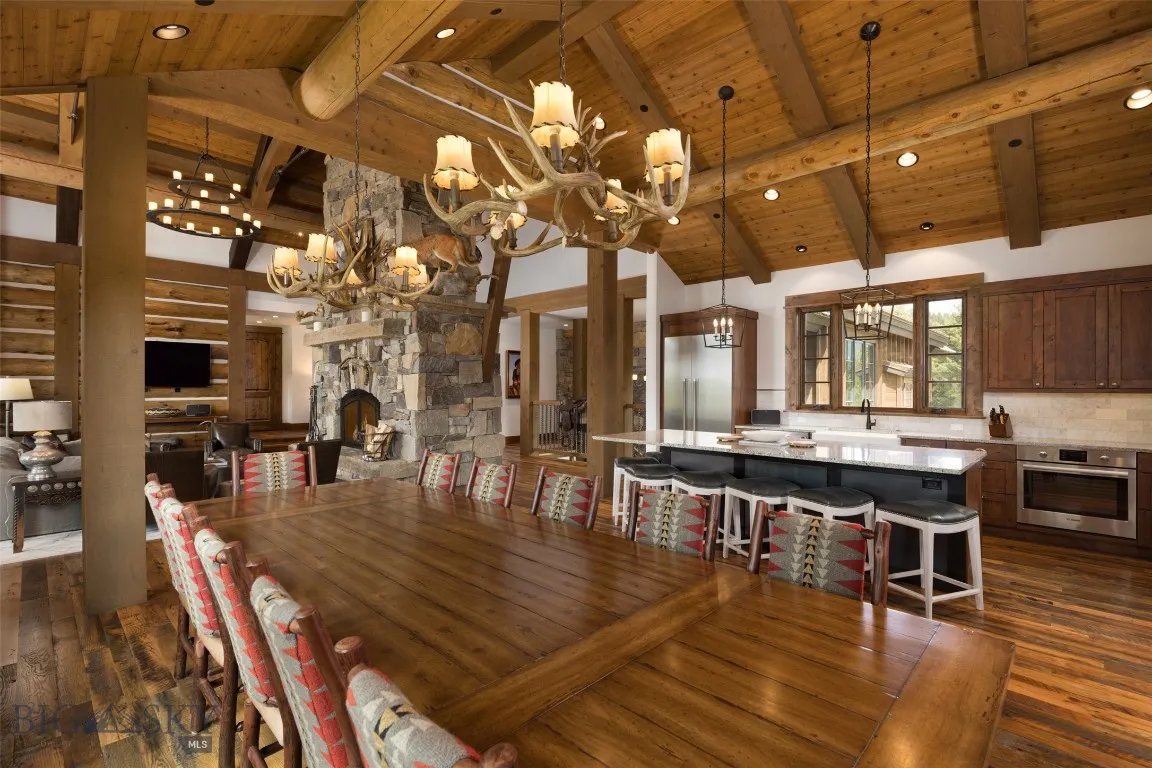 Spacious open-concept dining and living area featuring vaulted wood ceilings, a grand stone fireplace, and seating for large gatherings—perfect for entertaining in rustic luxury.