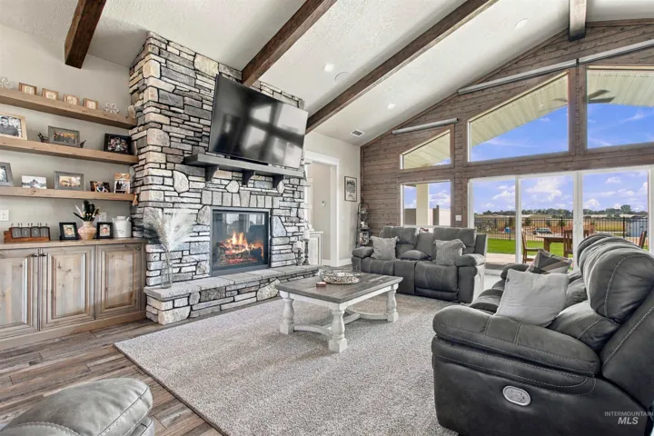 Vaulted ceiling with exposed wood beams, stone-clad fireplace with hearth, wood-finish flooring, extensive built-in shelving, and large windows with exterior access