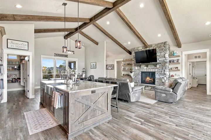Vaulted ceilings with exposed wood beams, stone fireplace, wood-finish flooring, kitchen island with built-in sink, and a walk-in pantry