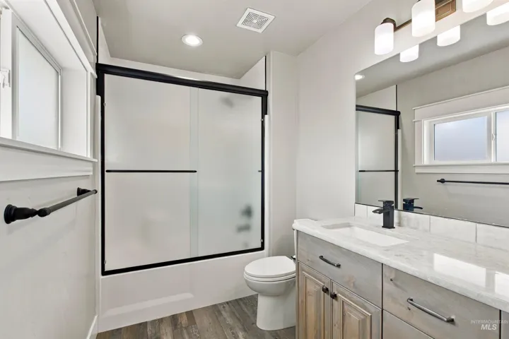 Bathroom featuring a vanity with a light-toned countertop, wood-finish flooring, a black-framed shower with frosted glass doors, a wall-mounted light fixture, and a large mirror