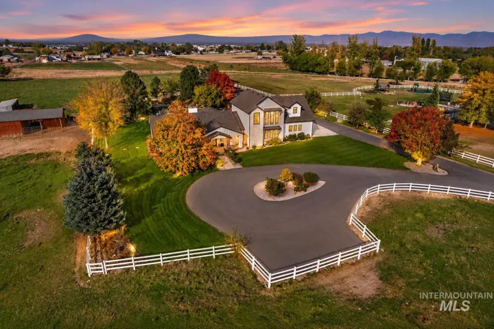 Two-story residence featuring a circular asphalt driveway, manicured landscaping, multi-rail perimeter fencing, and a detached outbuilding, set against a mountainous horizon