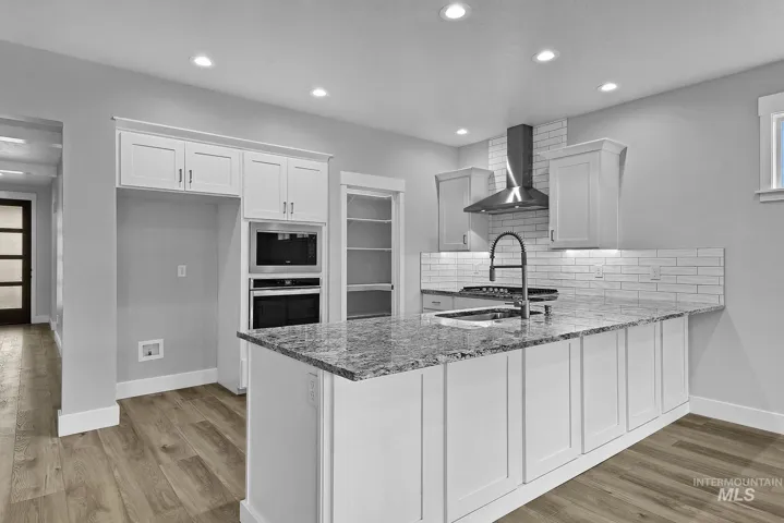 Kitchen featuring an expansive stone-surface island, white shaker-style cabinetry, a built-in microwave and wall oven, stainless steel range hood, and wood-finish flooring