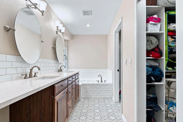 Dual vanity with integrated sinks and brushed nickel fixtures, white subway tile backsplash, white soaking tub with subway tile surround, patterned floor tile, and built-in closet shelving