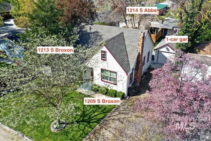 Elevated view showing the main home at front with additional units behind. Lawn, mature trees, and a flowering tree are visible. A driveway runs along the right side to a detached garage at the rear.