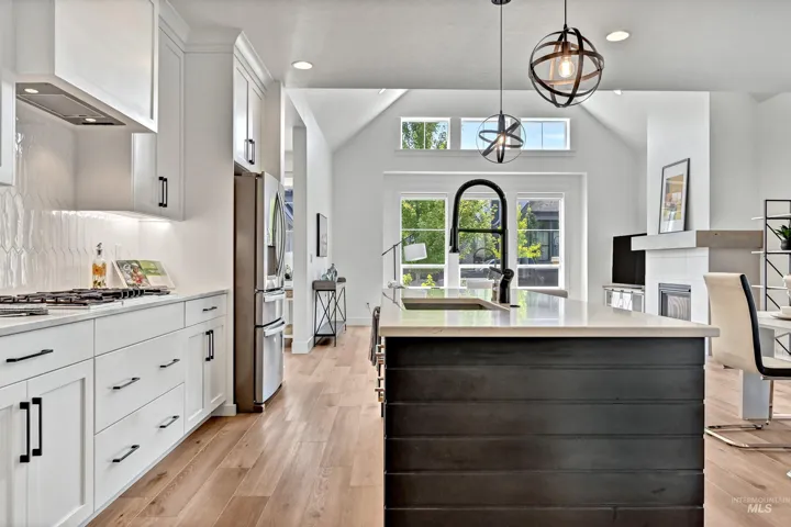 Modern kitchen featuring a central island with a sink and matte black faucet, light wood-finish flooring, white cabinetry with black hardware, and an integrated stainless steel refrigerator