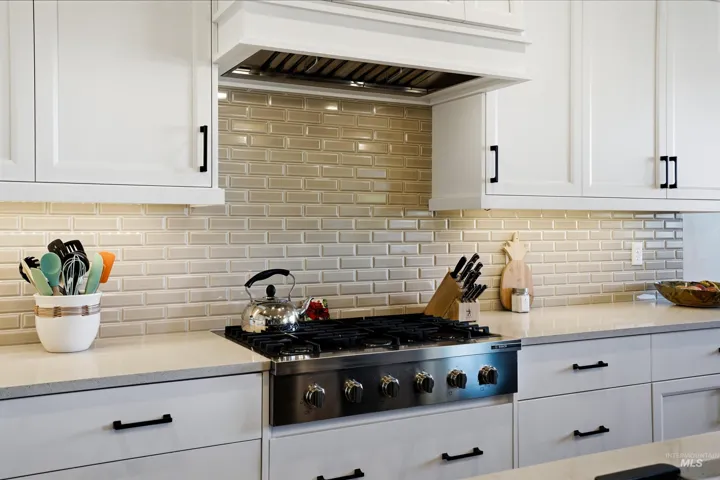 Kitchen featuring white Shaker-style cabinetry, matte black hardware, a light-toned stone-surface countertop, a glossy brick-patterned tile backsplash, and a stainless steel gas range with a built-in vent hood