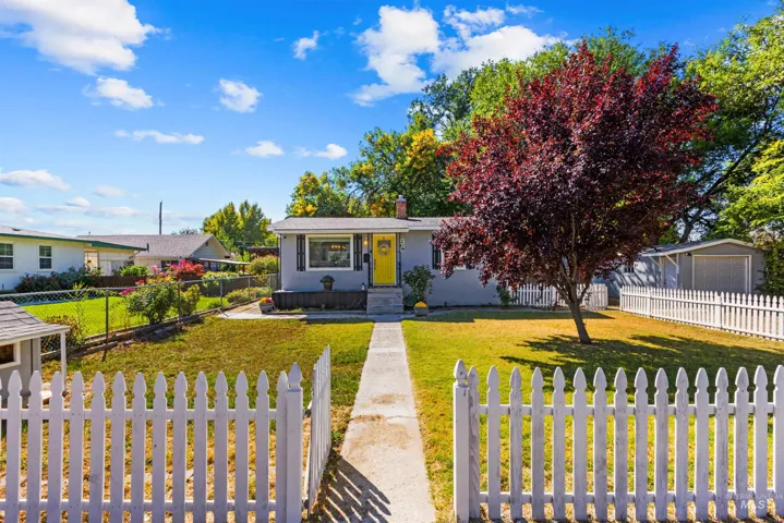 Property exterior featuring a white picket fence, mature tree, manicured lawn, and a light-colored façade with a yellow front door